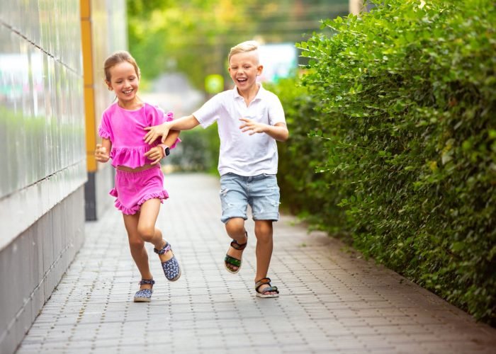 Two smiling kids, boy and girl running together in town, city in summer day. Concept of childhood, happiness, sincere emotions, carefree lifestyle. Little caucasian models in bright clothes.